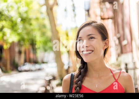 Happy asian woman relaxing enjoying weekend walk in her residential neighborhood. Sunny summer day serne girl walking in quiet street of modern condo buildings in urban city background. Stock Photo