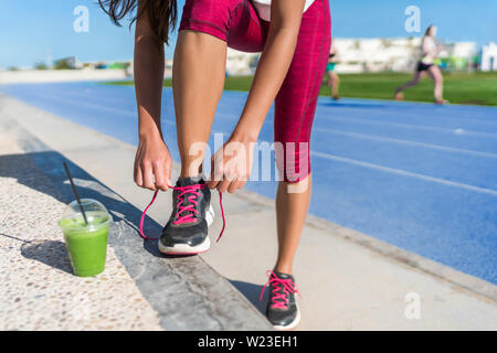 Healthy lifestyle woman runner tying running shoes drinking green smoothie cup juice drink before race workout on run tracks at outdoor stadium. Athlete getting ready for cardio training. Stock Photo