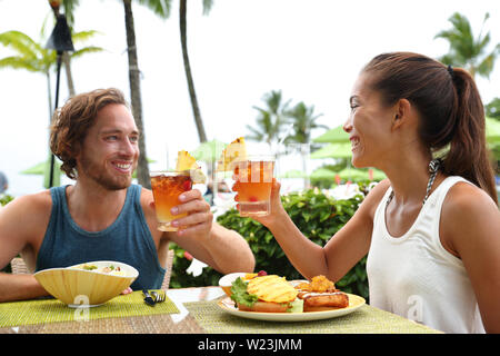 Happy multiracial couple toasting cheers with alcoholic hawaiian drinks, mai tai, Hawaii experience. Summer travel holidays, people enjoying local food meal at outdoor terrace restaurant of resort. Stock Photo