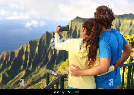 Couple on travel destination vacation relaxing together. Happy young adults tourists taking pictures with smartphone of amazing scenic view of na pali coast at Kalalau lookout on Kauai, Hawaii, USA. Stock Photo