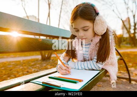 Little beautiful artist drawing with colored pencils, girl sitting on a bench in sunny autumn park, golden hour Stock Photo