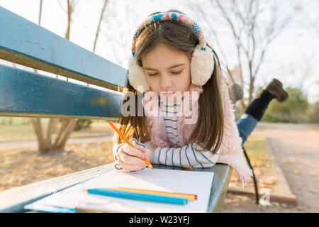 Little beautiful artist drawing with colored pencils, girl sitting on a bench in sunny autumn park, golden hour Stock Photo