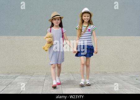 Two little beautiful girlfriends holding hands, girls walking in striped dresses, hats with backpack, background gray wall Stock Photo
