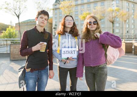 Group teenagers boy and two girls, with a notepad with handwritten word start. Teenagers looking forward, city background, golden hour Stock Photo