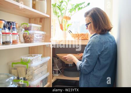 Interior of wooden pantry with products for cooking. Adult woman taking kitchenware and food from the shelves. Stock Photo