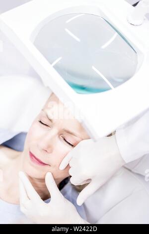 Beauty technician examining woman's facial skin using magnifying glass, close-up. Stock Photo