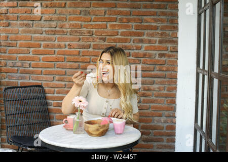 Blonde young female having morning breakfast with healthy food and pink cappuccino while sitting on a beautiful cozy garden with brick wall Stock Photo