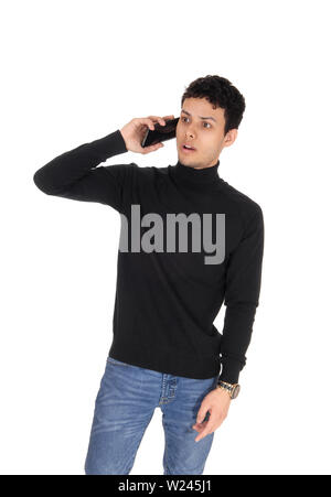 A young slim man in a black sweater and jeans standing, talking on the phone and is shocked, isolated for white background Stock Photo