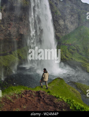 Gljufrafoss waterfall in gorge of mountains. Tourist attraction Iceland near falls of Seljalandsfoss. Man hiker in blue jacket standing on stone and looks at flow of falling water Stock Photo