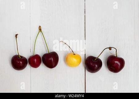 White cherry among red cherries on wooden background. Standing out from the crowd, individuality concept Stock Photo