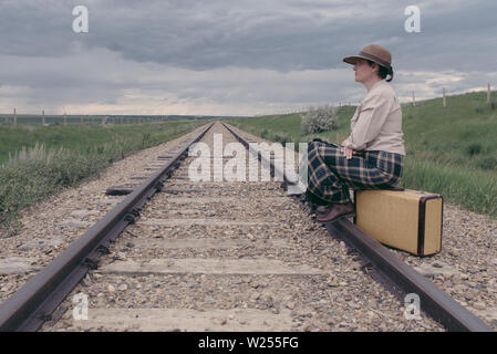 Young woman in historic dress sitting on vintage suitcase on railroad track looking ahead- journey concept Stock Photo