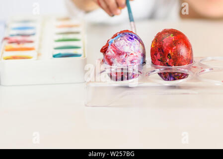 Child hands painted Easter eggs, paints and brushes on white table. Preparation for the Easter. Kid hands draw with colors. Stock Photo