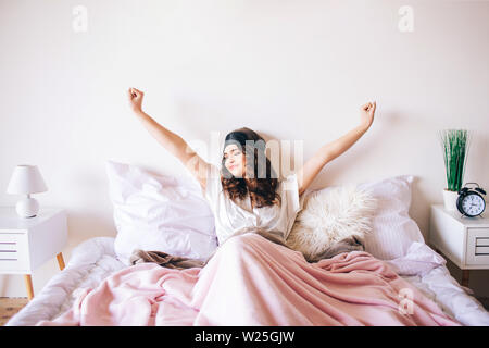 Dark haired beautiful young brunette wake up in her bed. Stretching up hands and smile. Sleeping mask on forehead. Alone in room. Bright daylight Stock Photo