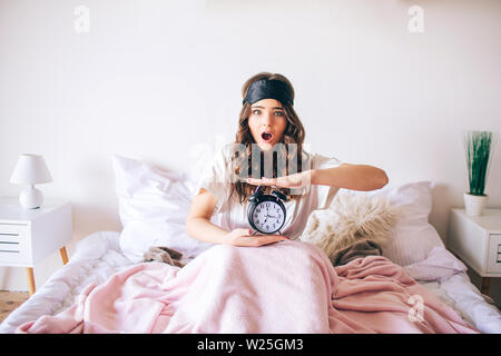 Dark haired beautiful young brunette wake up in her bed. Confused woman hold clock in hands. Woke up later. Amazed model look on camera with wonder. B Stock Photo