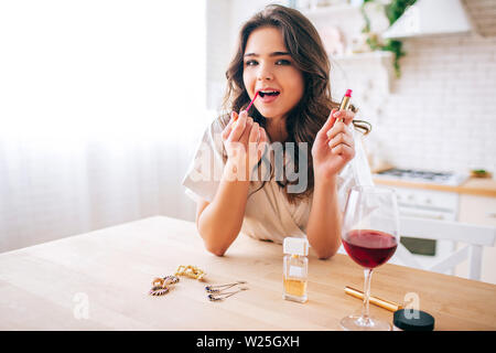 Young woman with dark hair standing in kitchen and putting make up on. Glss of red wine on table. Alone in kitchen. Wear morning gown Stock Photo