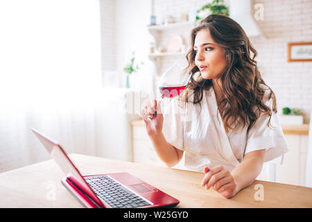 Young woman with dark hair standing in kitchen and drinking red wine. Enjoying time and life. Red laptop on table top. Alone in kitchen. Wearing morni Stock Photo