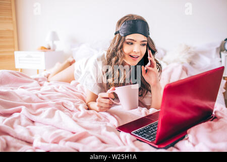 Young woman with dark hair. Talking on phone and looking on laptop screen. Cup in hand. Alone in bedroom. Lying on bed Stock Photo