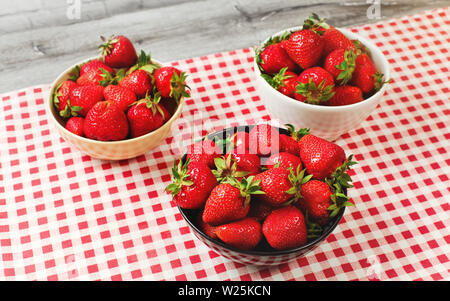 Three small bowls with ripe strawberries on desk with red gingham pattern tablecloth Stock Photo