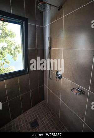 Modern bathroom in hotel room, steel shower, next to window with green leaves on tree behind Stock Photo