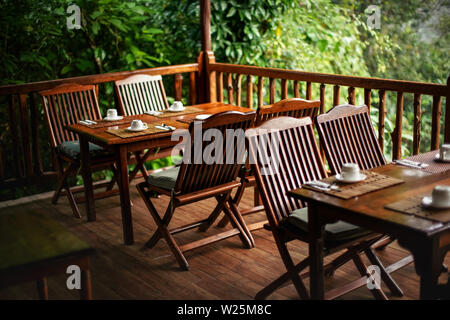 Empty wooden chairs and tables, with coffee cups upside down - ready for morning breakfast, jungle rainforest in background Stock Photo