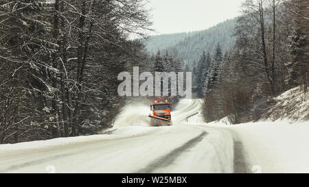 Bright orange plough truck cleaning snow on forest road in winter, view from car driving other way, trees around, overcast day Stock Photo