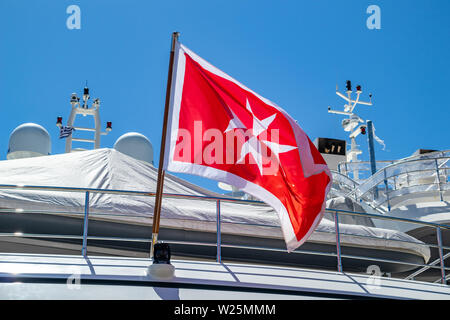 Maltese old flag and small greek flag on yacht stern. Boat at marina Zeas, Greece. Blue sky background, close up view. Stock Photo