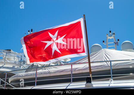 Maltese old flag and small greek flag on yacht stern. Boat at marina Zeas, Greece. Blue sky background, close up view. Stock Photo