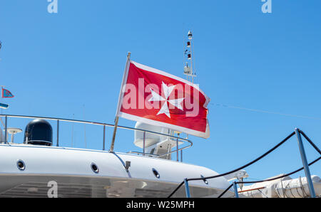 Maltese old flag and small greek flag on yacht stern. Boat at marina Zeas, Greece. Blue sky background, close up view. Stock Photo