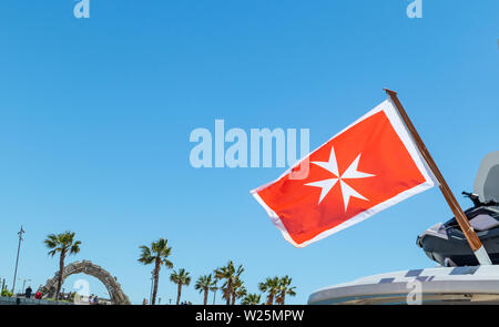 Maltese old flag and small greek flag on yacht stern. Boat at marina Zeas, Greece. Blue sky background, close up view. Stock Photo