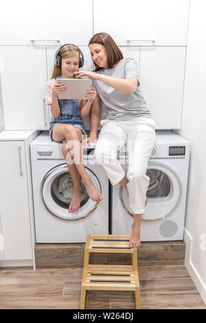 Young mother and her daughter are playing with tablet in laundry room sitting on washing machine Stock Photo