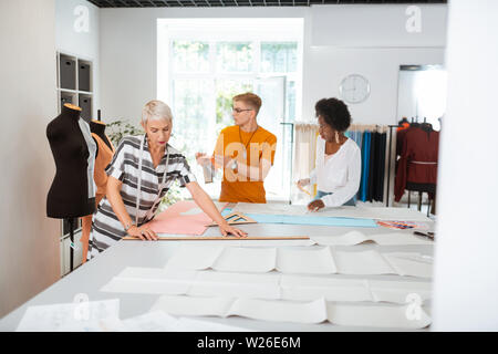Busy tailor. Serious aged female designer using a ruler while making a paper pattern on the cutting table Stock Photo