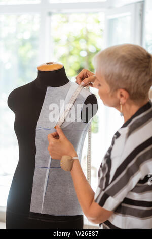 Dressmakers dummy. Short-haired blond female tailor standing with a pinball on her hand while taking measurements Stock Photo