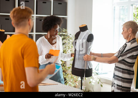 Motivated colleagues. Young female tailor holding a tablet while looking at her senior coworker taking measurements on the dummy Stock Photo