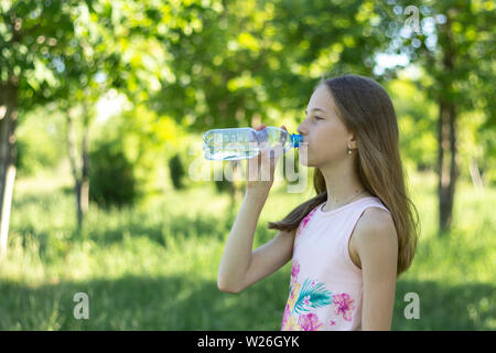 A young blonde girl in a pink t-shirt drinks water from a plastic bottle on a background of green forest. Young girl drinks water Stock Photo