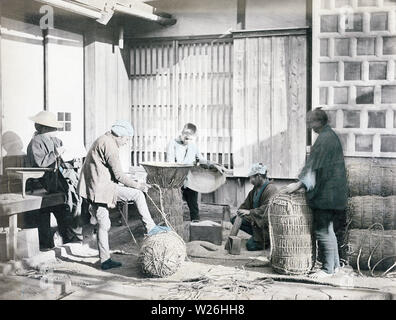 [ 1880s Japan - Packing Rice ] —   Two men in the foreground are packing rice in straw bags, while the man on the left is taking records. A soroban (Japanese abacus) lies on the table next to him. In the back a boy is filling a still open bag with rice. A masu wooden measuring box lies on the ground. Masu existed in many sizes from 0.9 to 18 liters.  19th century vintage albumen photograph. Stock Photo