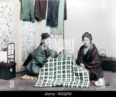 [ 1890s Japan - Japanese Women Sitting at a Table ] —   Two women in kimono and traditional hairstyles are sitting at a low table in a studio. One is reading a book. On the left is an ando lamp  19th century vintage albumen photograph. Stock Photo