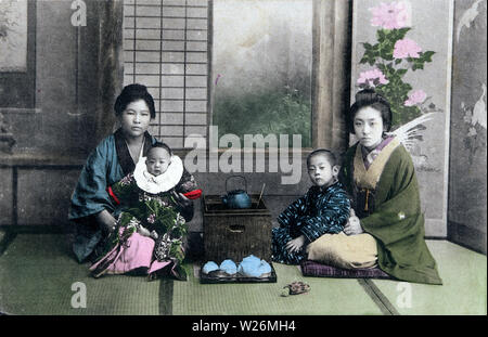 [ 1910s Japan - Japanese Mothers and Children ] —   Two mothers in kimono and traditional hairstyles and their children. They are seated next to a hakohibachi, a brazier encased in a wooden box. On the floor is a tray with a teapot and cups.  20th century vintage postcard. Stock Photo