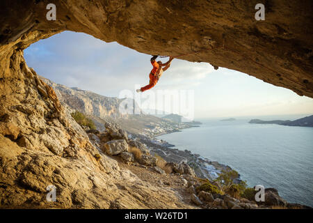 Young man climbing challenging route in cave against beautiful view of coast Stock Photo