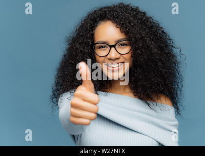 Portrait of dark skinned female has thumb up, shows agreement or approval, has smile on face, wears spectacles and jumper, gestures indoor against blu Stock Photo