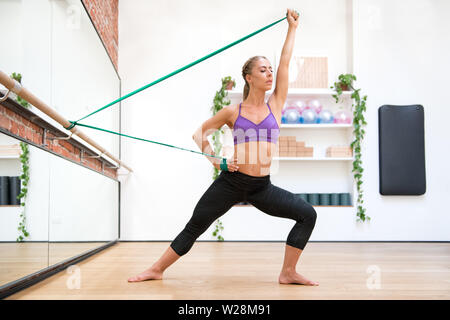 Young athlete woman doing arms workout with flex band in side lunge at gym Stock Photo
