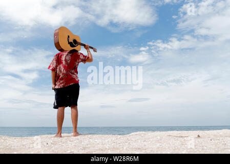 handsome young man holding guitar over shoulder on the beach with the blue sky Stock Photo
