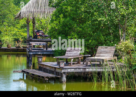 Two old wooden deck chairs near the lake water in a tropical garden, asia, Thailand. Travel and nature concept Stock Photo