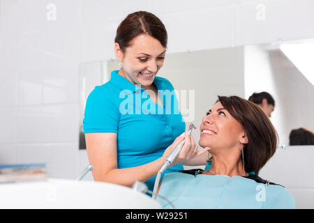 Dentist finishing successful teeth restoration, black haired doctor, in green mask, giving to happy patient mirror, beautiful long-haired woman Stock Photo