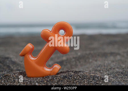 Close up picture of a small orange anchor beach toy placed on the black sand at the Batu Bolong beach in Canggu, Bali during a cloudy day Stock Photo