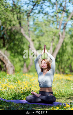 Image of young woman doing yoga with arms raised sitting in lotus position on blue rug in forest