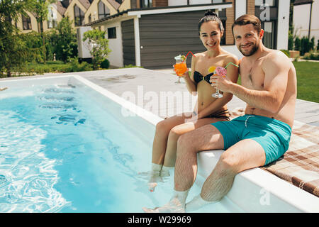 Lovely heteroseksual couple rented a house with a swimming pool. Man and woman sitting near water pool and holding fresh cocktail Stock Photo