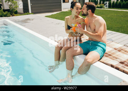 Lovely heteroseksual couple having summer resting. Man and woman sitting near water pool and holding fresh cocktail Stock Photo