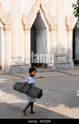 Bagan, Myanmar - March 2019: Child carrying rolled bamboo mat in Ananda temple backyard in Bagan