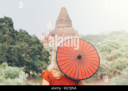 Blonde Caucasian woman with red traditional umbrella looks towards Ananda temple and pagodas of ancient Bagan in Myanmar Stock Photo