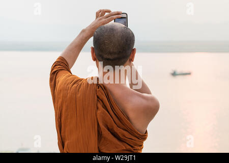 Bagan, Myanmar - March 2019: Burmese Buddhist monk in orange robe taking pictures of Irrawaddy river at sunset.